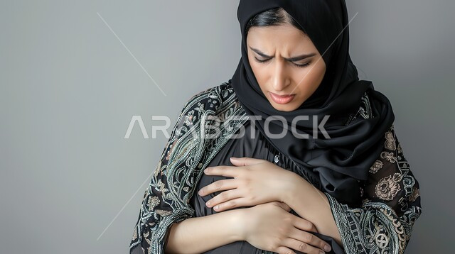 الشعور بالشعور بآلام المخاض  وقرب الولاده، حرقة في المعدة وعسر الهضم، الإرهاق والتعب والإحساس بالمغص، بورتريه مقرب لإمرأة عربية سعودية خليجية محجبة حامل ترتدي العباءة تمسك بطنها بإيماءات الشعور بالألم، الحاجة لوضع خطة علاجية، خلفية رمادية