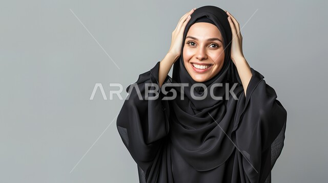 السرور والسعادة لسماع الأخبار السارة، الدهشة لحدوث أمر ما، تعابير المفاجأة والذهول, بورتريه مقرب لإمرأة عربية سعودية خليجية محجبة مبتسمة تضع يديها على رأسها بايماءات الاستغراب, خلفية رمادية 
