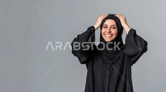 تعابير المفاجأة والذهول, الإستغراب بحدوث أمر ما، سماع الأخبار السارة والشعور بالسعادة والسرور، بورتريه مقرب لإمرأة عربية سعودية خليجية محجبة مبتسمة تضع يديها على رأسها بايماءات الدهشة, خلفية رمادية 