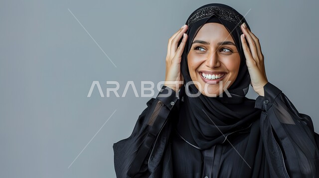 الإستغراب بحدوث أمر ما، سماع الأخبار السارة والشعور بالسعادة والسرور، إيماءات المفاجأة والذهول, بورتريه مقرب لإمرأة عربية سعودية خليجية محجبة مبتسمة تضع يديها على رأسها بتعابير الدهشة, خلفية رمادية 