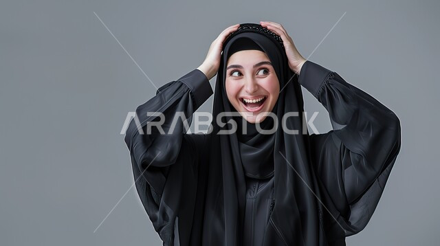 الشعور بالسعادة والسرور بسماع الأخبار السارة، إيماءات المفاجأة والذهول, بورتريه مقرب لإمرأة عربية سعودية خليجية محجبة مبتسمة تضع يديها على رأسها بتعابير الدهشة, الإستغراب بحدوث أمر ما، خلفية رمادية 