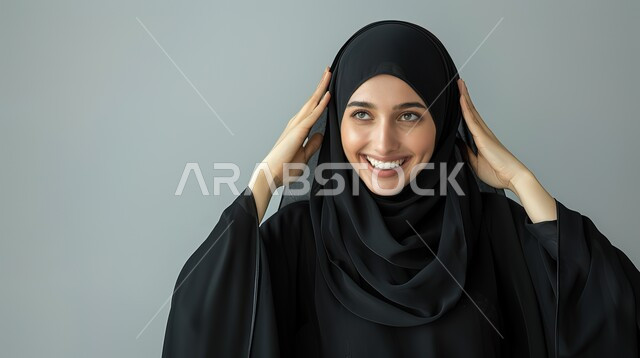إيماءات المفاجأة والذهول, الشعور بالسعادة والسرور بسماع الأخبار السارة، بورتريه مقرب لإمرأة عربية سعودية خليجية محجبة مبتسمة تضع يديها على رأسها بتعابير الدهشة, الإستغراب بحدوث أمر ما، خلفية رمادية 