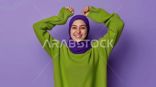 النجاح وتحقيق الأهداف، مفهوم الفرح بالنصر والفوز، تعابير السرور والسعادة، بورتريه مقرب لامرأة عربية خليجية سعودية محجبة مبتسمة ترتدي الزي الكاجوال ترفع يديها بإيماءات الحماس، خلفية بنفسجية