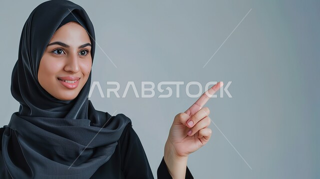 تحديد موقع معين بإيماءات البشاشة، إشارات وحركات يدين تدل على هذا المكان (هنا)، بورتريه مقرب لامرأة عربيه خليجيه سعوديه مبتسمه محجبه ترفع اصابعها السبابة وتشير الى شيء ما، خلفية رمادية