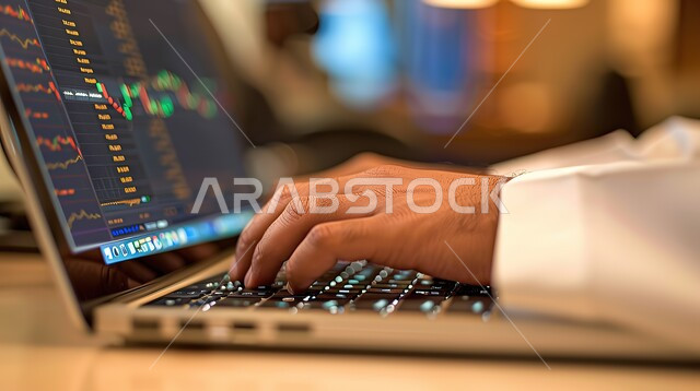 متابعة البورصة من خلال استخدام جهاز الكمبيوتر المحمول, العمل في التجارة الالكترونية, صورة مقربة ليد رجل عربي خليجي سعودي  في مكان عمله في الشركة، الرسوم البيانية، صعود وهبوط البورصة المالية، الاستثمار المالي والاقتصادي، مجال المال والأعمال