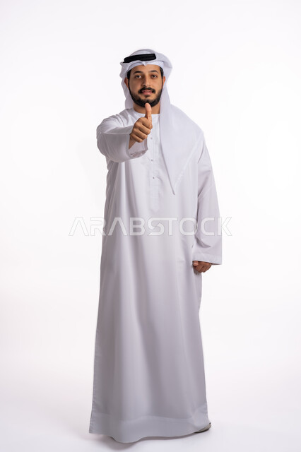 مفهوم الجودة والامتياز، بورتريه لرجل عربي خليجي إماراتي يرتدي الكندورة الإماراتية مع الغترة البيضاء يرفع يده بإيماءات القبول والموافقة، النظر إلى الكاميرا بتعابير الإعجاب والرضا، صورة لكامل الجسم، خلفيه بيضاء