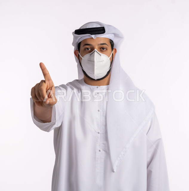 إشارات وحركات يدين تدل على هذا المكان (هنا)، تجنب الملامسه السطحيه والحفاظ على المسافات، إتباع الإجراءات الوقائيه ضد فايروس كورونا، بورتريه مقرب لرجل عربي خليجي اماراتي يرتدي الكندورة والغتره يضع القناع الطبي يشير باصبعه إلى الأعلى، تحديد موقع شيء معين، خلفيه بيضاء