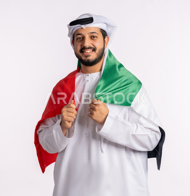 مفهوم الاعتزاز والفخر بإنجازات البلاد، هوية اليوم الوطني الاماراتي 51، الإحتفال باليوم الوطني الإماراتي، احياء ذكرى استقلال الإمارات، بورتريه مقرب لرجل عربي خليجي إماراتي يرتدي الكندورة والغترة البيضاء يقوم بحمل علم الإمارات العربية المتحدة على ظهره، الحب والانتماء للوطن، خلفية بيضاء
