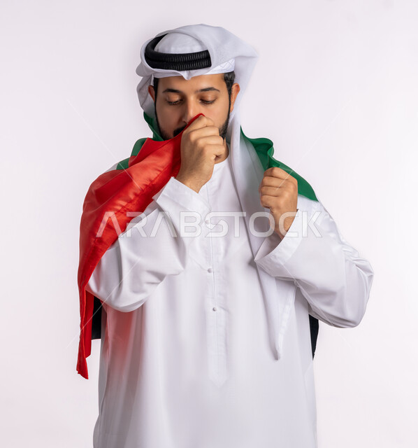 الاعتزاز والفخر بإنجازات البلاد، هوية اليوم الوطني الاماراتي 51، الإحتفال باليوم الوطني الإماراتي، يوم استقلال الإمارات، الحب والانتماء للوطن، بورتريه مقرب لرجل عربي خليجي إماراتي يرتدي الكندورة والغترة البيضاء يقوم بتقبيل علم الإمارات العربية المتحدة، خلفية بيضاء