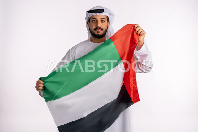 الاعتزاز والفخر بإنجازات البلاد، هوية اليوم الوطني الاماراتي 51، الإحتفال باليوم الوطني الإماراتي، يوم استقلال الإمارات، الحب والانتماء للوطن، بورتريه مقرب لرجل عربي خليجي إماراتي يرتدي الكندورة والغترة البيضاء يقوم برفع علم الإمارات العربية المتحدة، خلفية بيضاء