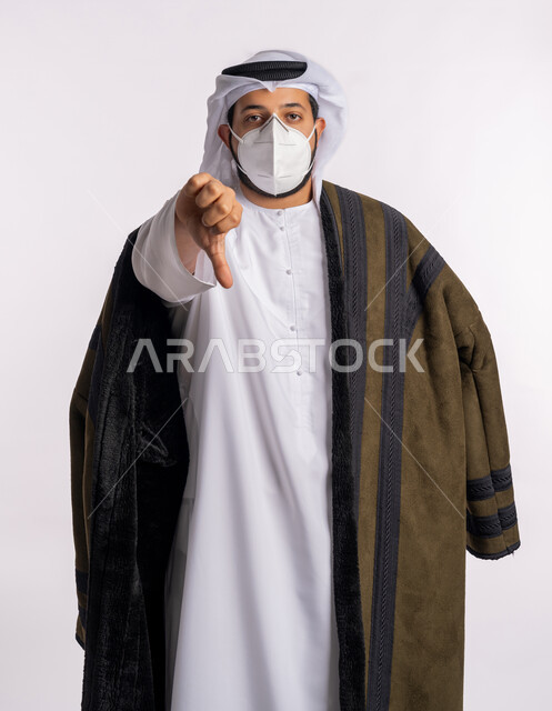 الشعور بالإنزعاج من شيء ما، مفهوم الرفض والفشل، تجنب الملامسه السطحيه والحفاظ على المسافات، إتباع الإجراءات الوقائيه ضد فايروس كورونا، بورتريه لرجل عربي خليجي اماراتي يرتدي الفروة والغتره يضع القناع الطبي يخفض ابهامه بايماءات الرفض وعدم القبول، خلفيه بيضاء