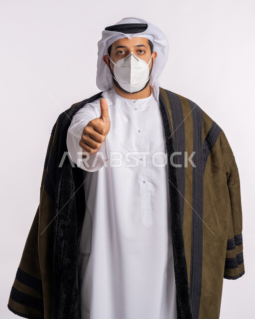 مفهوم الجودة والامتياز، بورتريه لرجل عربي خليجي إماراتي يرتدي الفروة الغترة البيضاء يضع لكمامة الطبية يرفع يده بإيماءات القبول والموافقة، إتباع الإجراءات الوقائيه ضد فايروس كورونا، تعابير الإعجاب والرضا، جنب الملامسه السطحيه والحفاظ على المسافات، خلفية بيضاء