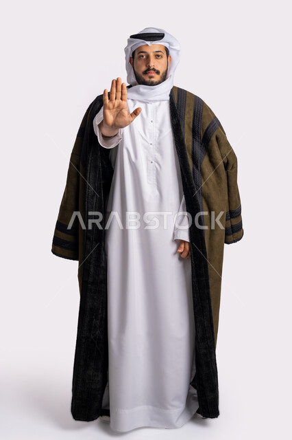 طلب المنع والتوقف عن فعل شيء ما، إيماءات الرفض والامتناع، بورتريه لرجل عربي خليجي إماراتي يرتدي الفروة والغترة ينظر إلى الكاميرا بتعابير الحزم والجدية، ارتداء الملابس المناسبة لفصل الشتاء، صورة لكامل الجسم، خلفية بيضاء