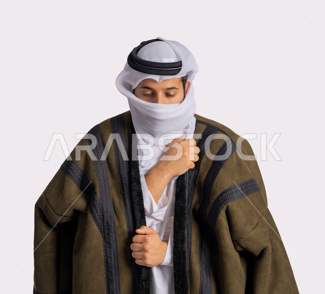 إيماءات وجه ويدين تدل على البرد الشديد، الأجواء الشتوية الباردة، الشعور بالصقيع والقشعريرة والتجمد، بورتريه مقرب لرجل عربي خليجي اماراتي يرتدي الفروة والغترة البيضاء يقوم بتغطية انفه وتدفئة يديه، خلفية بيضاء