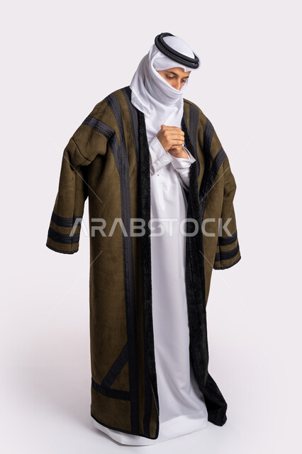 إيماءات وجه ويدين تدل على البرد الشديد، الأجواء الشتوية الباردة، الشعور بالصقيع والقشعريرة والتجمد، بورتريه لرجل عربي خليجي اماراتي يرتدي الفروة والغترة البيضاء يقوم بتغطية انفه وتدفئة يديه، صورة الجسم بالطول الكامل، خلفية بيضاء
