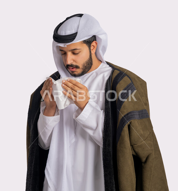 استعمال المناديل الورقية أثناء العطس، الشعور بالتعب والمرض، الإصابة بفايروس كورونا كوفيد19، بورتريه مقرب لرجل عربي خليجي اماراتي يرتدي الفروة والغترة البيضاء، الحاجة لتلقي العلاج، إيماءات وجه ويدين تدل على العطاس، خلفية بيضاء