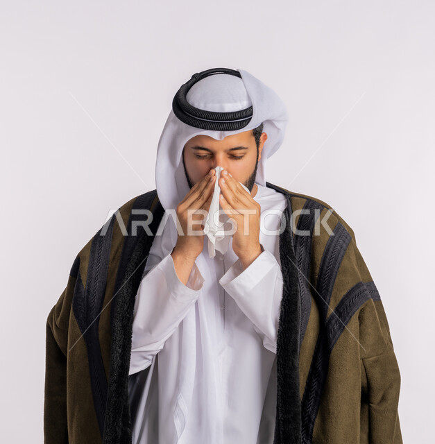 استعمال المناديل الورقية أثناء العطس، الشعور بالتعب والمرض، الإصابة بفايروس كورونا كوفيد19، بورتريه مقرب لرجل عربي خليجي اماراتي يرتدي الفروة والغترة البيضاء، الحاجة لتلقي العلاج، إيماءات وجه ويدين تدل على العطاس، خلفية بيضاء