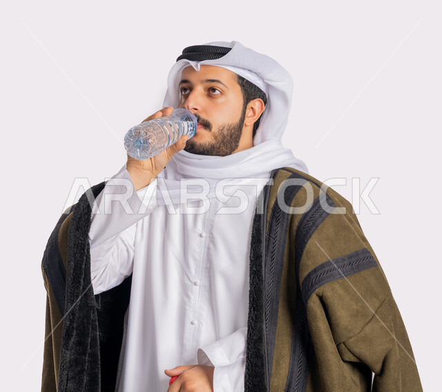 Feeling thirsty and dehydrated, a close-up portrait of an Emirati Gulf Arab man wearing a white fur and ghutra, drinking water from a small plastic bottle, maintaining the health of the body’s immune system, continuing to drink water, cold winter weather, white background