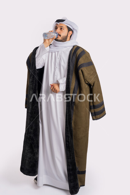 المداومة على شرب المياه، المحافظة على سلامة الجهاز المناعي للجسم، الشعور بالعطش والجفاف، بورتريه لرجل عربي خليجي اماراتي يرتدي الفروة والغترة البيضاء يقوم بشرب الماء من زجاجة بلاستيكية صغيرة، الأجواء الشتوية الباردة، صورة الجسم بالطول الكامل، خلفية بيضاء