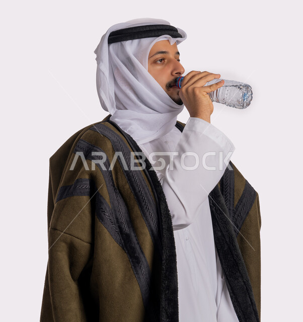 المحافظة على سلامة الجهاز المناعي للجسم، الشعور بالعطش والجفاف، بورتريه مقرب من الجانب لرجل عربي خليجي اماراتي يرتدي الفروة والغترة البيضاء يقوم بشرب الماء من زجاجة بلاستيكية صغيرة، المداومة على شرب المياه، الأجواء الشتوية الباردة، خلفية بيضاء