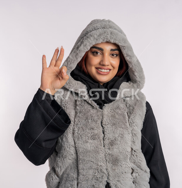 The concept of concession and quality, expressions of admiration and contentment, a close portrait of an Emirati Gulf Arab woman wearing a winter jacket that raises her hand with the gestures of acceptance and approval, a Saudi young woman looking at the camera with expressions of pleasure and happiness, cold winter atmosphere, gray background