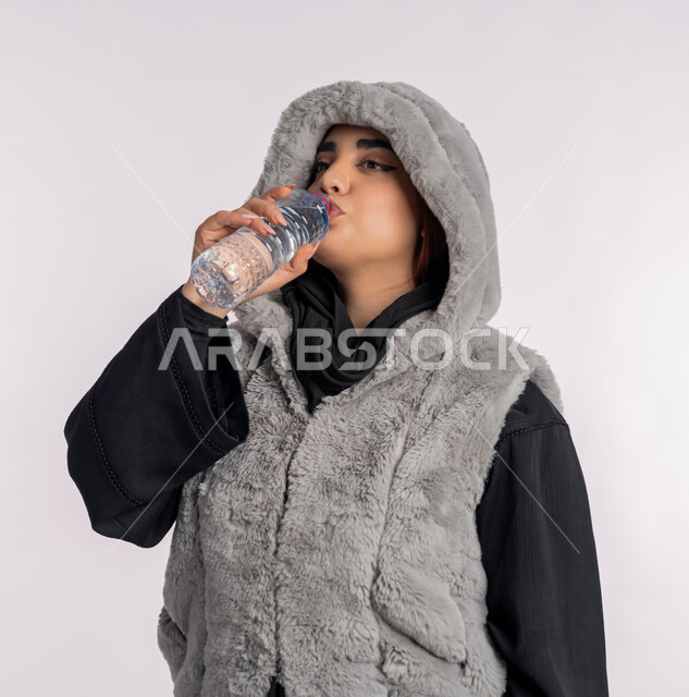 الشعور بالعطش والجفاف، بورتريه مقرب لامرأة عربية خليجيه اماراتية ترتدي معطف شتوي، شابة سعودية تقوم بشرب الماء من زجاجة بلاستيكية صغيرة، المحافظة على سلامة الجهاز المناعي للجسم، المداومة على شرب المياه، الأجواء الشتوية الباردة، خلفية رمادية