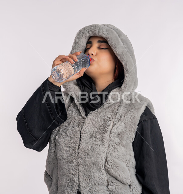 المداومة على شرب المياه، الأجواء الشتوية الباردة، الشعور بالعطش والجفاف، بورتريه مقرب لامرأة عربية خليجيه اماراتية ترتدي معطف شتوي، شابة سعودية تقوم بشرب الماء من زجاجة بلاستيكية صغيرة، المحافظة على سلامة الجهاز المناعي للجسم، خلفية رمادية