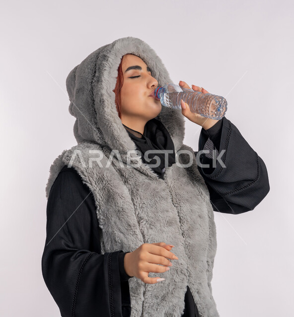 المحافظة على سلامة الجهاز المناعي للجسم، المداومة على شرب المياه، الأجواء الشتوية الباردة، الشعور بالعطش والجفاف، بورتريه مقرب من الجانب لامرأة عربية خليجيه اماراتية ترتدي معطف شتوي، شابة سعودية تقوم بشرب الماء من زجاجة بلاستيكية صغيرة، خلفية رمادية