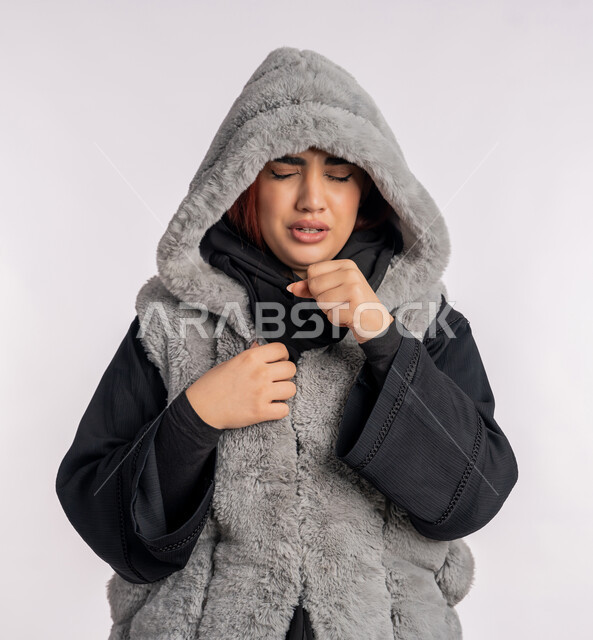 الاصابة بفايروس كورونا كوفيد19، الاجواء الشتوية الباردة، الانفلونزا والتهاب الرئة، بورتريه مقرب لامرأة عربية خليجيه اماراتية ترتدي معطف شتوي تعاني من السعال، شابة سعودية تشعر بالمرض والكحة، خلفية رمادية