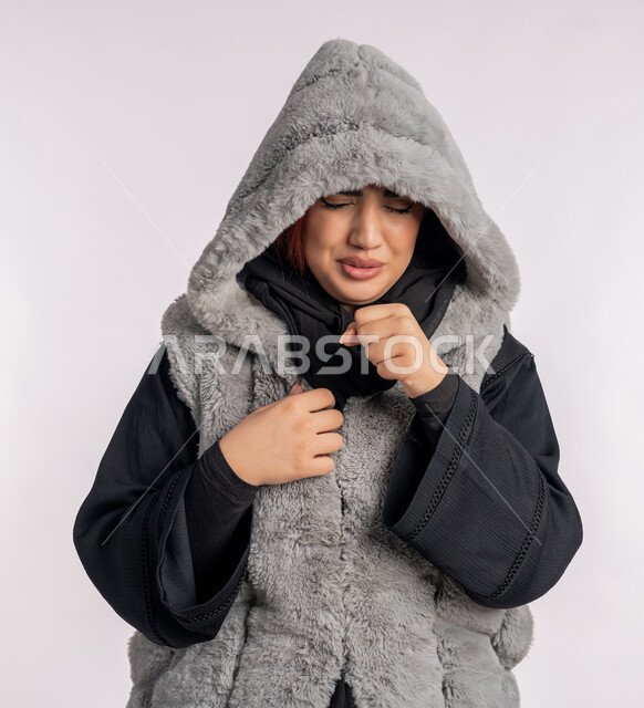 الاجواء الشتوية الباردة، الشعور بالمرض والكحة، الانفلونزا والتهاب الرئة، بورتريه مقرب لامرأة عربية خليجيه اماراتية ترتدي معطف شتوي تعاني من السعال، شابة سعودية مصابة بفايروس كورونا كوفيد19، خلفية رمادية