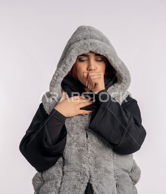 Influenza and lung inflammation, a feeling of disease and cough, cold winter atmosphere, a close portrait for an Emirati Gulf Arab woman wearing a winter coat suffering from coughing, a young Saudi woman with virus Corona Kovid 19, gray background