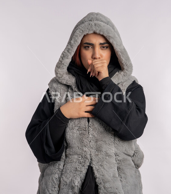 الشعور بالمرض والكحة، الانفلونزا والتهاب الرئة، الاجواء الشتوية الباردة، بورتريه مقرب لامرأة عربية خليجيه اماراتية ترتدي معطف شتوي تعاني من السعال، شابة سعودية مصابة بفايروس كورونا كوفيد19، خلفية رمادية