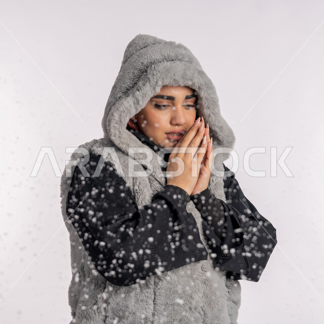 الاستمتاع بالأجواء الشتوية الثلجية الباردة، بورتريه مقرب من الجانب لامرأة عربية خليجيه اماراتية ترتدي معطف شتوي تقوم بجمع يديها بجانب وجهها، شابة سعودية تنظر بإيماءات تدل على الاحساس بالبرد الشديد، موسم تساقط الثلج، الشعور بالصقيع والقشعريرة والتجمد، خلفية رمادية