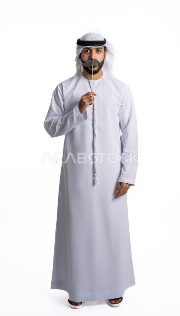 يوم الرجل 19 نوفمبر , بورتريه لرجل عربي إماراتي خليجي  يرتدي الكندورة والغترة البيضاء يحمل بيده عود خشبي  مع قصاصة ورقية على شكل شارب يضعه على فمه, ينظر إلى الكاميرا بإيماءات الحزم والجدية, الصحة الجسدية للذكور,   الطول الكامل للجسم, خلفية بيضاء