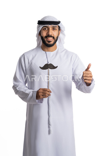 الإشارة بإصبع الابهام نحو الأعلى بإيماءات الجودة والإعجاب بشيء ما, ذكرى اليوم العالمي للرجال 19 نوفمبر , بورتريه لرجل عربي إماراتي خليجي يرتدي الكندورة والغترة البيضاء يحمل بيده  قصاصة ورقية على شكل شارب  ينظر إلى الكاميرا بتعابير السرور,الاهتمام بصحة الذكور, خلفية بيضاء