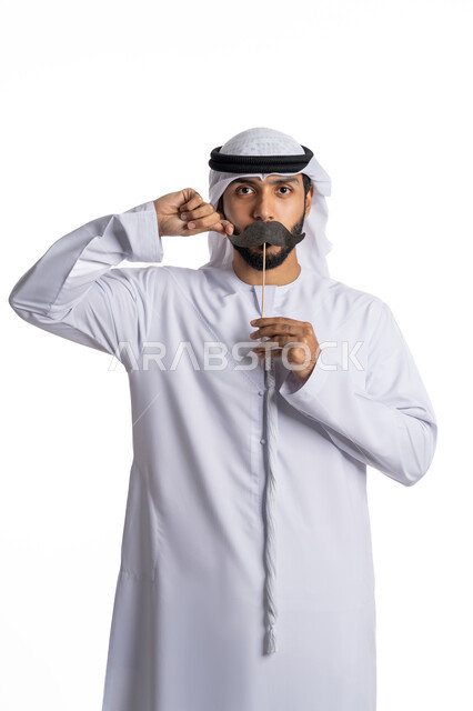 International Men's Day, November 19, annual global events, a portrait of an Arab Gulf Emirati man wearing a kandura and a ghutra and holding a black mustache-shaped piece of paper in his hand, standing straight and looking at the camera and pointing to the piece of paper, the concept of caring for a man, white background