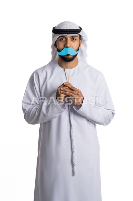 الاحفال بالمناسبات العالمية السنوية، اليوم العالمي للرجل 19 نوفمبر، بورتريه لشاب عربي خليجي إماراتي يرتدي الغترة والكندورة ويقف باستقامة وينظر إلى الكاميرا ويمسك بيديه قصاصة ورقية على شكل شارب ،مفهوم الاهتمام بالرجل، خلفية بيضاء