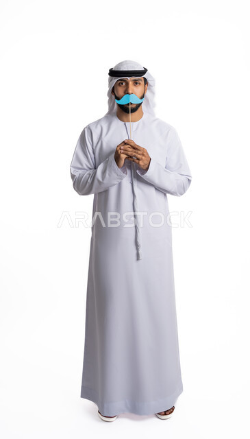 الاحتفال  بمناسبات عالمية، بورتريه لرجل عربي خليجي إماراتي يرتدي الغترة والكندورة ويقف بشكل مستقيم يمسك بيديه قصاصة ورقية باللون الأزرق على شكل شارب وينظر إلى الكاميرا بإيماءات الحزم والجدية، الاهتمام بصحة الرجل، الطول الكامل للجسم، خلفية بيضاء
