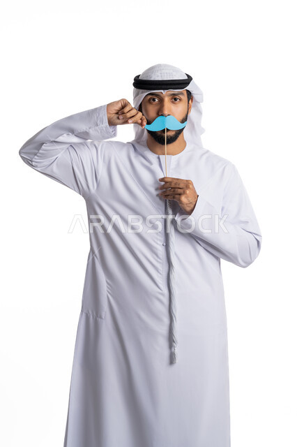 الاحتفال باليوم العالمي للرجل 19 نوفمبر، بورتريه لرجل عربي خليجي إماراتي يرتدي الكندورة والغترة ويحمل بيده قصاصة ورقية على شكل شارب، الوقوف باستقامة والنظر إلى الكاميرا والإشارة إلى القصاصة، مفهوم الاهتمام بالرجل، خلفية بيضاء