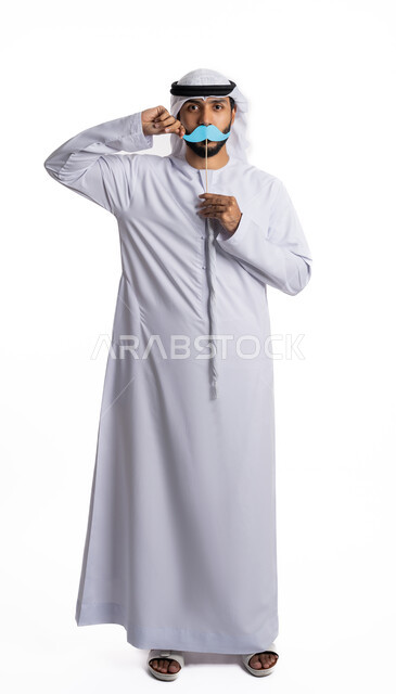 الاحتفال السنوي باليوم العالمي للرجل 19 نوفمبر، بورتريه لرجل عربي خليجي إماراتي يرتدي الكندورة والغترة ويحمل بيده قصاصة ورقية على شكل شارب باللون الأزرق، الوقوف باستقامة والنظر إلى الكاميرا والإشارة إلى الورقة، مفهوم الاهتمام بالرجل، الطول الكامل للجسم، خلفية بيضاء
