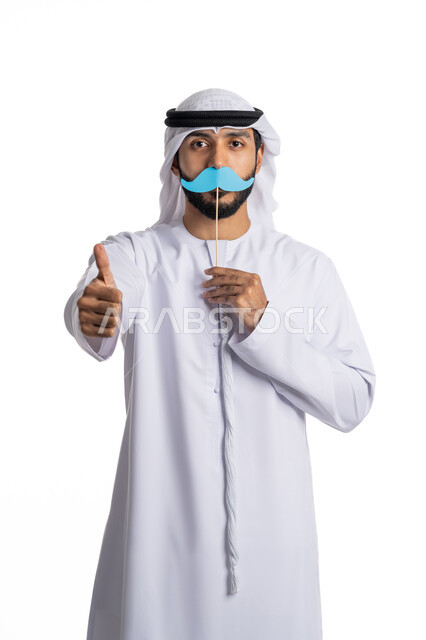 ذكرى اليوم العالمي للرجل 19نوفمبر، بورتريه لرجل عربي خليجي إماراتي يرتدي الغترة والكندور يحمل بيده قصاصة ورقية باللون الأزرق على شكل شارب وينظر إلى الكاميرا بتعابير السرور، الإشارة باصبع الإبهام بإيماءات الإعجاب والقبول، الطول الكامل للجسم، خلفية بيضاء