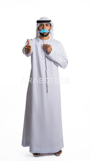 ذكرى اليوم العالمي للرجل 19نوفمبر، بورتريه لرجل عربي خليجي إماراتي يرتدي الغترة والكندور يحمل بيده قصاصة ورقية باللون الأزرق على شكل شارب وينظر إلى الكاميرا بتعابير السرور، الإشارة باصبع الإبهام بإيماءات الإعجاب والقبول، الطول الكامل للجسم، خلفية بيضاء