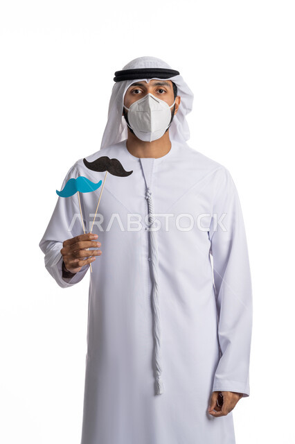 الاهتمام بالصحة العامة، الاجراءات المتخذة للحماية من كوفيد 19, بورتريه لشاب عربي خليجي إماراتي يرتدي الثوب الإماراتي التقليدي ويضع الكمامة للوقاية من فايروس كورونا ويحمل بيده قصاصات ورقية على شكل شارب, مناسبات عالمية, خلفية بيضاء