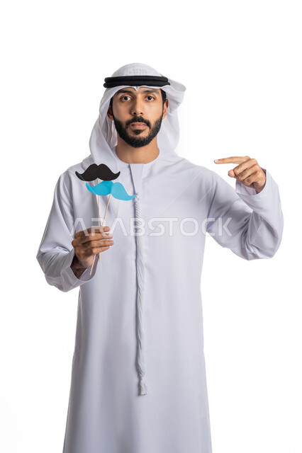 الاحتفال السنوي باليوم العالمي للرجل 19 نوفمبر، بورتريه لرجل عربي خليجي إماراتي يرتدي الكندورة والغترة يحمل بيده قصاصات ورقية على شكل شارب باللون الأزرق والأسود, وينظر إلى الكاميرا، الإشارة باصبع السبابة إلى  الورقة بإيماءات الفخر والثقة بالنفس، مفهوم الاهتمام بالرجل، مناسبات عالمية سنوية، خلفية بيضاء