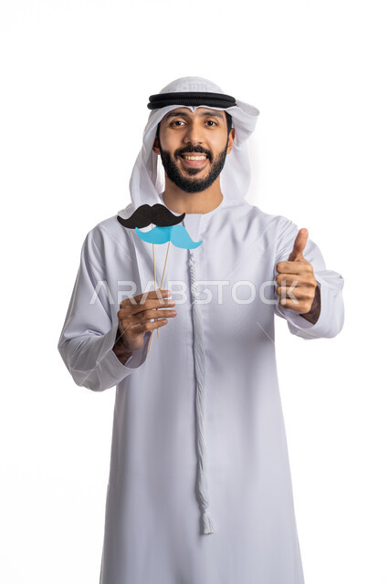ايماءات البهجة والسرور، رفع اليد بعلامات القبول والإعجاب، الاحتفال باليوم العالمي للرجل 19 نوفمبر، بورتريه لشاب عربي خليجي إماراتي يرتدي الكندورة والغترة يحمل بيده قصاصات ورقية على شكل شارب باللون الأزرق والأسود، مفهوم الاهتمام بالرجال، خلفية بيضاء