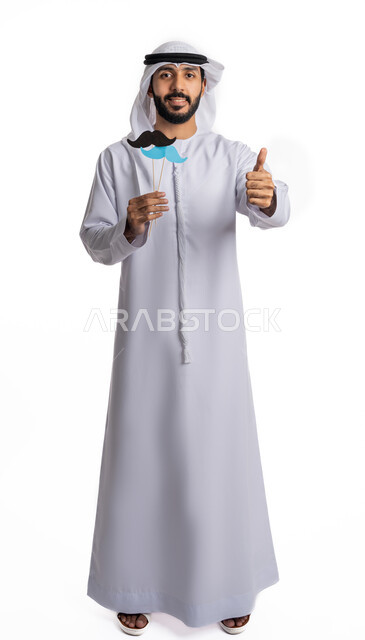 رفع اليد بعلامات القبول والإعجاب، الاحتفال باليوم العالمي للرجل 19 نوفمبر، بورتريه لرجل عربي خليجي إماراتي يرتدي الكندورة والغترة يحمل بيده قصاصات ورقية على شكل شارب باللون الأزرق والأسود، مفهوم الاهتمام بالرجال، صورة لكامل الجسم، خلفية بيضاء