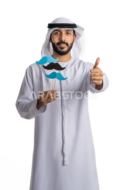 The concept of strength and masculinity, looking at the camera with expressions of satisfaction, raising the hand with signs of acceptance and admiration, celebrating International Men’s Day November 19, portrait of an Arab Gulf Emirati young man wearing a kandora and ghutra, holding in his hand paper clips in the shape of a mustache in blue and black, white background.