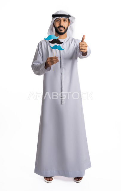 حمل قصاصات ورقية على شكل شارب باللون الأزرق والأسود، مفهوم النظر إلى الكاميرا بتعابير الرضا، الاحتفال باليوم العالمي للرجل 19 نوفمبر، بورتريه لشاب عربي خليجي إماراتي يرتدي الكندورة والغترة يرفع يده بعلامات القبول والإعجاب، صورة لكامل الجسم، خلفية بيضاء
