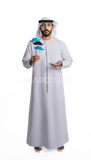 رفع اليد بإيماءات عرض شيء ما، الاهتمام بالمظهر الخارجي، الاحتفال باليوم العالمي للرجل، احترام وتقدير الرجال، بورتريه لشاب عربي خليجي إماراتي يرتدي الكندورة والغترة يمسك بعود خشبي يحمل قصاصات ورقية على شكل شارب، صورة لكامل الجسم، خلفية بيضاء