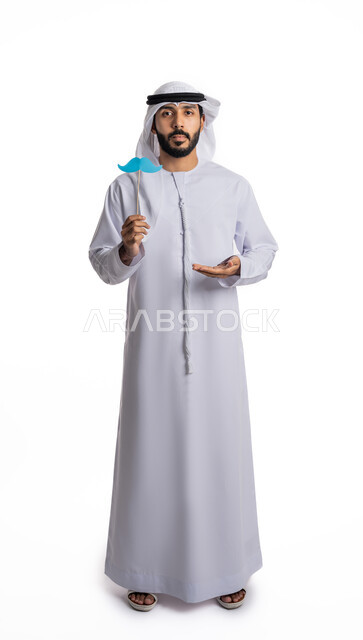 الاهتمام بالمظهر الخارجي، الاحتفال باليوم العالمي للرجل، احترام وتقدير الرجال، بورتريه لشاب عربي خليجي إماراتي يرتدي الكندورة والغترة يمسك بعود خشبي يحمل قصاصة ورقية على شكل شارب يرفع يده بإيماءات عرض شيء ما، صورة لكامل الجسم، خلفية بيضاء
